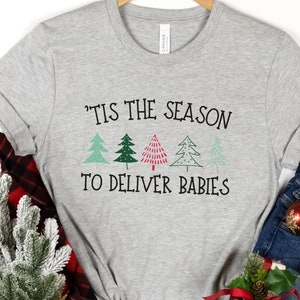 May include: A gray t-shirt with the text "'Tis the season to deliver babies" and a graphic of three stylized Christmas trees. The shirt is on a white background with a red and black plaid blanket, a small green tree, and blue jeans with red ornaments.
