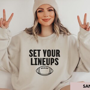 May include: A beige sweatshirt with the text "SET YOUR LINEUPS" and a black outline of a football. The sweatshirt is being modelled by a person wearing a beige beanie.