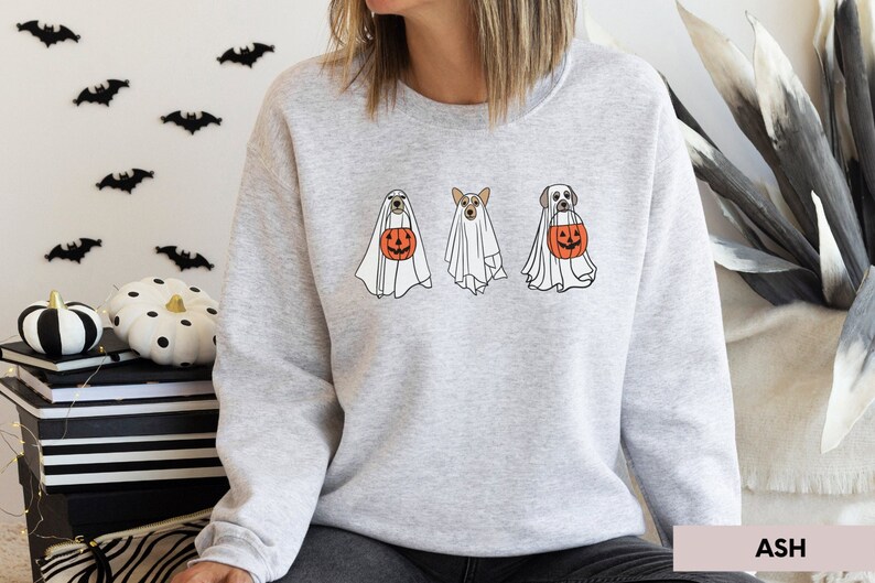 Peut inclure: Un sweat-shirt gris avec un motif de trois chiens habill&eacute;s en fant&ocirc;mes tenant des citrouilles. Le texte "ASH" est visible dans le coin inf&eacute;rieur droit de l'image.