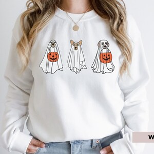 Peut inclure: Sweat-shirt blanc avec un motif de trois chiens habill&eacute;s en fant&ocirc;mes tenant des citrouilles. Le texte "WHITE" est visible dans le coin inf&eacute;rieur droit de l'image.