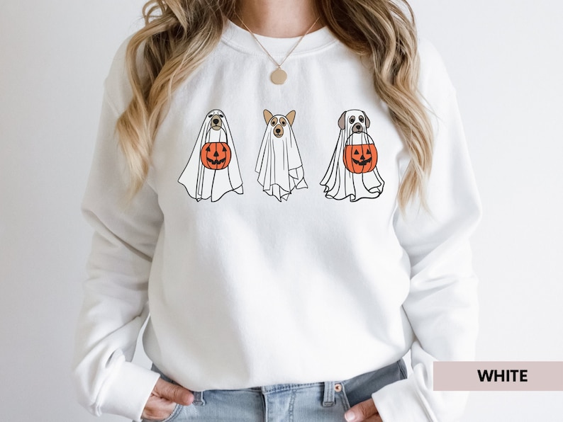 Peut inclure: Sweat-shirt blanc avec un motif de trois chiens habill&eacute;s en fant&ocirc;mes tenant des citrouilles. Le texte "WHITE" est visible dans le coin inf&eacute;rieur droit de l'image.