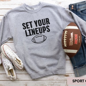 May include: A grey sweatshirt with the text "SET YOUR LINEUPS" and a black outline of a football. The sweatshirt is lying on a white wooden surface with a pair of white trainers and a brown football.