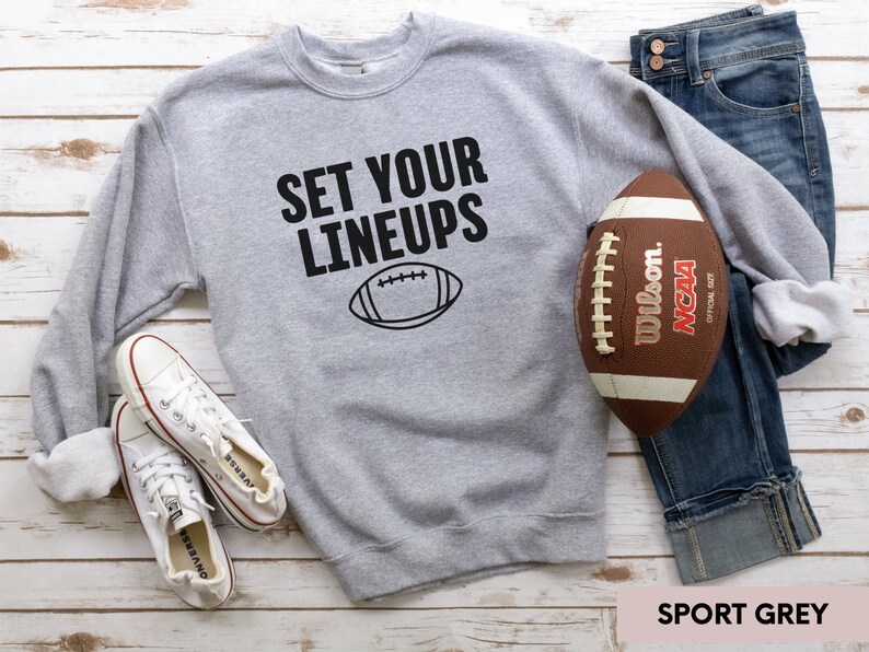 May include: A grey sweatshirt with the text "SET YOUR LINEUPS" and a black outline of a football. The sweatshirt is lying on a white wooden surface with a pair of white trainers and a brown football.