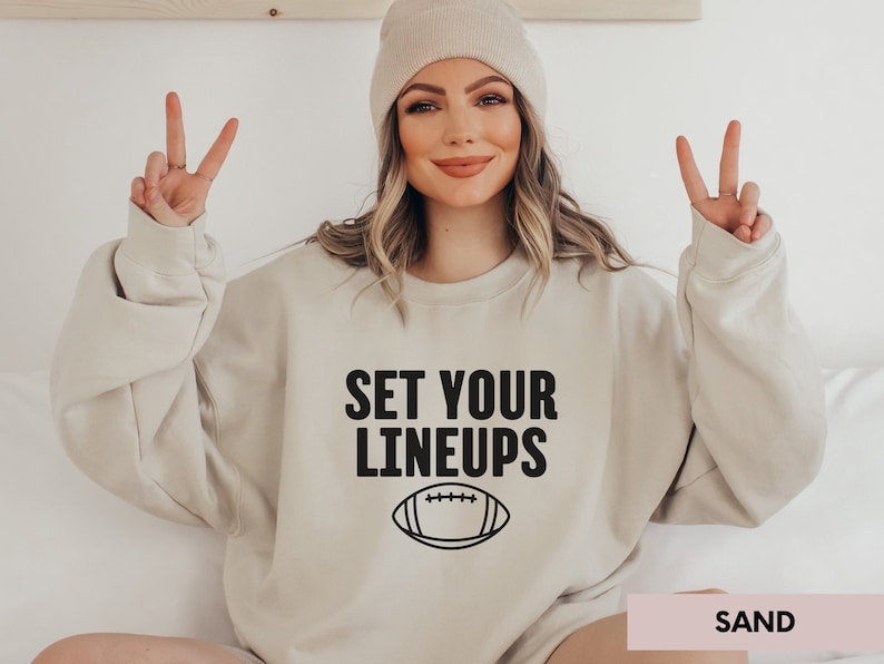 May include: A beige sweatshirt with the text "SET YOUR LINEUPS" and a black outline of a football. The sweatshirt is being modelled by a person wearing a beige beanie.