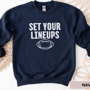 May include: Navy blue sweatshirt with the text "SET YOUR LINEUPS" and a white outline of a football.