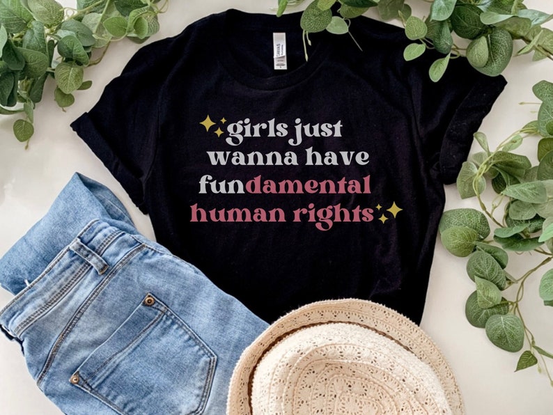 Puede incluir: Camiseta negra con el texto "girls just wanna have fundamental human rights" en blanco y rosa. La camiseta se combina con vaqueros azules y un sombrero de paja, con follaje verde en el fondo.