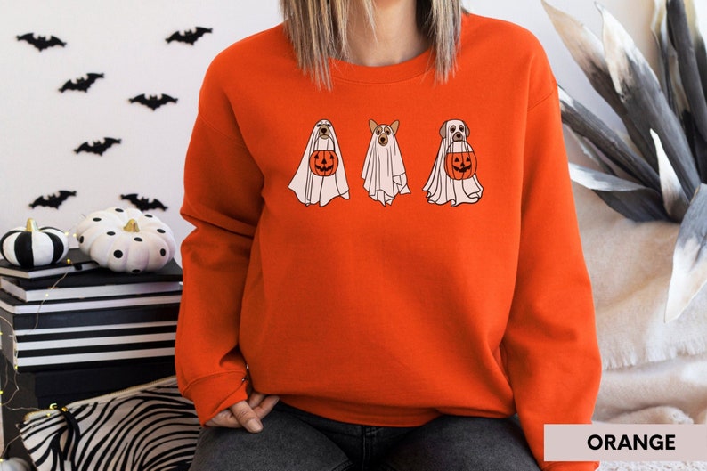 Peut inclure: Sweat-shirt orange avec un motif de trois chiens habill&eacute;s en fant&ocirc;mes tenant des citrouilles. Les chiens portent des costumes de fant&ocirc;mes blancs avec la t&ecirc;te qui d&eacute;passe. Le texte "ORANGE" est dans le coin inf&eacute;rieur droit de l'image.