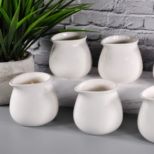 May include: Set of six white ceramic creamer pitchers with a rounded shape. The pitchers are arranged in two rows of three on a gray surface.