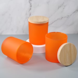 May include: Three cylindrical orange glass jars with a frosted finish. One jar is lying on its side, one stands upright with a light-colored wooden lid, and one stands upright without a lid, accompanied by a separate matching wooden lid nearby.