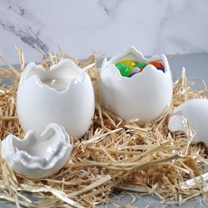 May include: Four white ceramic egg-shaped candle holders, two with the top half removed, one with colorful candy inside, and one with a white candle inside. The holders are sitting on a bed of straw.