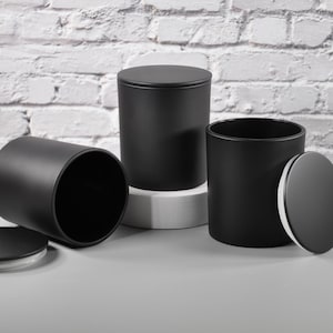 May include: Three matte black cylindrical candle jars with matching lids. One jar is standing upright with its lid on, one is lying on its side with the lid nearby, and one is standing open with the lid placed vertically against its side.