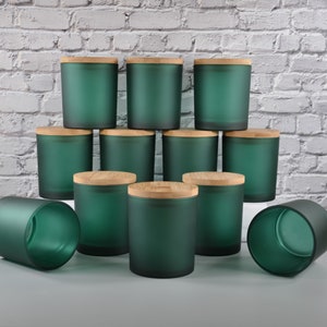 May include: A collection of eleven cylindrical candle jars made of frosted dark green glass. Nine of the jars are upright and topped with circular light-colored wooden lids, while two jars are positioned on their sides without lids.