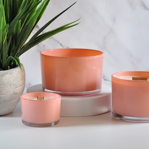 May include: Three pink glass candle jars with wooden wicks. The candles are of varying sizes and are arranged on a white surface.
