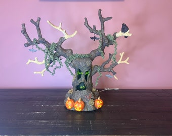 Op de afbeelding: Een Halloween-boomdecoratie met een griezelig gezicht, gloeiende groene ogen en vleermuizen die aan de takken hangen. De boom is versierd met oranje pompoenen en heeft een bruine, getextureerde schors.