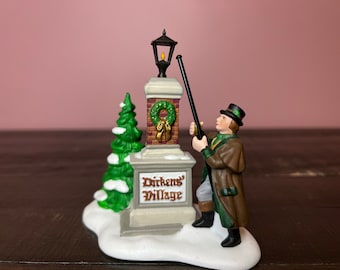 May include: A miniature Dickens Village figurine depicting a lamplighter with a long pole, a lamp post, and a Christmas tree. The lamp post has a wreath and the words "Dickens Village". The figurine is set on a snowy base.