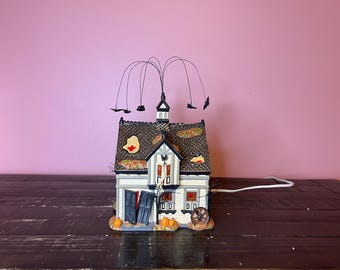 Op de afbeelding: Een miniatuurhuisje met een donker dak van dakspanen met herfstbladaccenten, een witte buitenkant en blauwe luiken. De structuur is bekroond met dunne draadtakken met kleine zwarte vleermuissilhouetten en bevat oranje pompoenen.