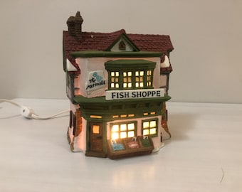May include: A ceramic miniature of a two-story fish shop with a red tile roof and green trim. The shop has a sign that reads "FISH SHOPPE" and "The Mermaid". The windows are lit, and a display case shows fish.