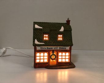 May include: A miniature ceramic bakery with a green shingled roof and snow accents. The sign reads "White Horse Bakery" with a horse silhouette. The windows glow with warm light, and a small wreath hangs on the door. A white electrical cord is attached.