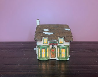 May include: A white ceramic cottage with a brown thatched roof and green trim. The cottage has a sign that reads "Cottage Toys" and a lit window. The cottage is sitting on a dark wooden surface.