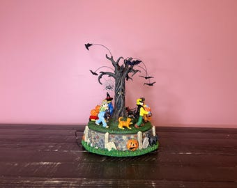 Op de afbeelding: Een Halloween-muziekspeelgoed met een spookachtige boom, vleermuizen en kinderen in kostuums. De kinderen dragen pompoenen en lopen naar de boom toe. De basis van het speelgoed is versierd met pompoenen, bladeren en een stenen muur.
