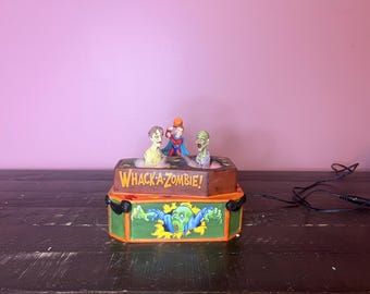 Op de afbeelding: Een houten doos met een "WHACK-A-ZOMBIE!" bord aan de voorkant. De doos heeft drie zombiekoppen die eruit steken. De doos staat op een donkerbruin oppervlak.