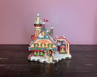 Puede incluir: Una miniatura de la estación de esquí Elf Mountain con techo verde, chimenea de piedra y un teleférico rojo. La estructura cuenta con ventanas iluminadas, una bandera roja con un copo de nieve y detalles nevados en la base y el techo.