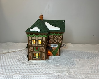 Puede incluir: Una casa iluminada de un pueblo navideño con tejado verde y detalles de nieve blanca. La casa tiene una fachada marrón y beige con un letrero que dice "T. Puddlewick". Las ventanas están iluminadas y una corona adorna la entrada.