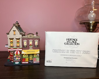 May include: A handpainted porcelain Johnson's Grocery & Deli building from the Heritage Village Collection Christmas in the City series, accompanied by its original white box featuring the collection name and product details in black text.