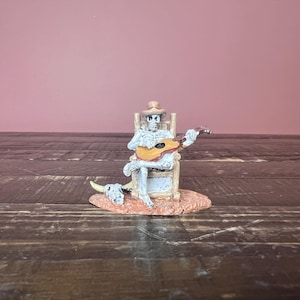 May include: A figurine of a skeleton playing a guitar while seated in a chair. The skeleton wears a hat and is accompanied by a cow skull. The figurine is painted in shades of white, brown, and orange.