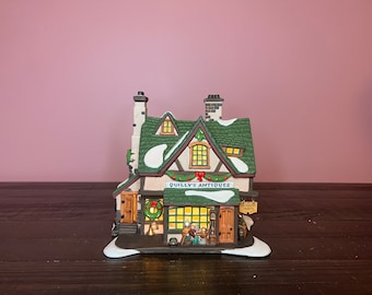 May include: A detailed ceramic model of "Quilly's Antiques", featuring a green roof with snow, and a lit interior. The shop has a wreath and a sign. It is set on a dark wooden surface, with a pink background.