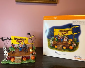 Op de afbeelding: Een Halloween-dorpsscène met een podium, skeletten en pompoenen. Het podium heeft een gele banner met de tekst "Halloween Dance".