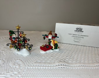Puede incluir: Dos figuras de porcelana de la Heritage Village Collection tituladas "Don't Break the Ornaments". Una representa a una figura decorando un árbol de metal negro con adornos coloridos, mientras que la otra muestra a dos figuras manipulando adornos.