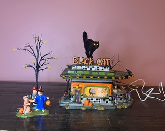 Op de afbeelding: Een Halloween-miniatuurgebouw genaamd Black Cat Diner met een zwart kattenfiguur op het dak, verlichte gele ogen en een bord met de tekst Black Cat Diner. Daarnaast staan een miniatuurboom en drie figuren in kostuums.