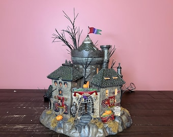 Op de afbeelding: Een gedetailleerd, meerlaags, spookachtig dorpsdecor met een stenen toren, een groen dak en een poort met een bord waarop "Haunted House Inn" staat. De scène is versierd met pompoenen, wijnstokken en een vlag met een schedel en gekruiste beenderen.