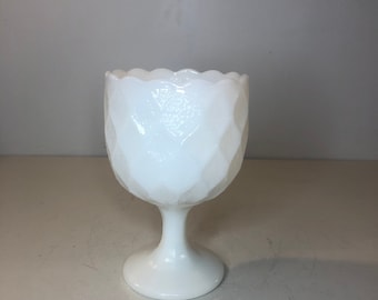 May include: A white milk glass pedestal bowl with a scalloped rim and a diamond pattern. The bowl is on a white surface.