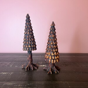May include: Two wooden pine tree sculptures with brown and gold paint. The trees are on a dark wood surface.