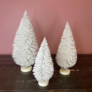 May include: Three white flocked artificial Christmas trees of varying sizes on a dark wood surface. The trees are all in a classic pine shape.