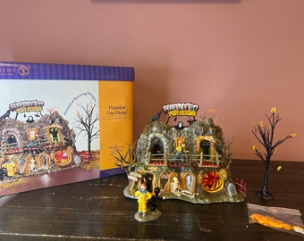 Op de afbeelding: Een miniatuur scène van een spookhuis met een bord met de tekst "Haunted Fun House". Het huis is gemaakt van steen en heeft een griezelig, fantasierijk ontwerp. Er staan ​​verschillende figuren voor het huis, waaronder een clown, een skelet en een zwarte kat.
