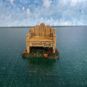 May include: A miniature wooden bench with the words "Harvest Time" on the front. The bench is light brown and sits on a patch of green grass with red and orange leaves. The background is a blurred blue and white.