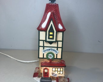 May include: A miniature light-up building titled Rimpy's Bakery with a tall red roof, white walls, and dark trim. The structure features a green wreath with an R, a red door, a sign reading Fresh Cookies, and white snow accents on the roof and base.