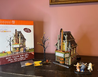 Op de afbeelding: Een Department 56 Snow Village Halloween LaGhosti Movie Theater cadeau-set inclusief een miniatuur bioscoopgebouw, een kale boom, een gele bank met een zwarte kat, een pompoen en drie kleine figuren in kostuums.