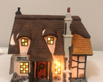 May include: A ceramic cottage with a thatched roof, white walls, and a lit window. The cottage has a sign that reads "Maylie Cottage" and a wreath on the door.