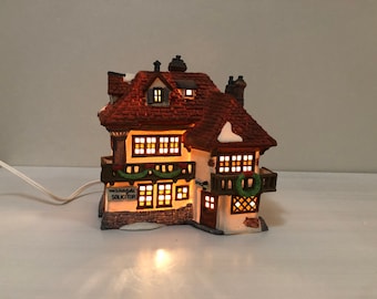 Puede incluir: Una casa en miniatura de cerámica con tejado de tejas rojas, ventanas iluminadas y un pequeño balcón. La casa tiene una fachada de piedra y una corona verde. El texto "Meadowfield Solicitor" es visible. Un cable eléctrico blanco está conectado.