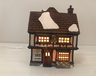 May include: A ceramic Christmas village house with a brown tiled roof and white snow detailing. The house features lit windows, a wreath, and the text "TUTBURY PRINTER". A power cable is visible, extending from the base.