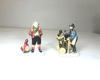 May include: Two miniature figurines. One is a shoemaker working on a shoe cart. The other figure wears a red jacket, black trousers, and pink socks. The figurines are on a white base. The scene is reminiscent of a Christmas market.
