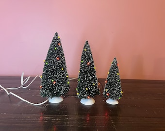 May include: Three artificial bottle brush Christmas trees of varying heights, featuring dark green needles with white tips, each mounted on a white circular base and decorated with small, multi-colored LED lights connected by a thin white wire.