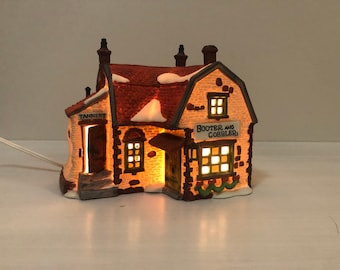 May include: A ceramic Christmas village house with a red tile roof and snow detailing. The building has illuminated windows and the text "Booter and Cobbler" and "Tannery". A power cable extends from the base.