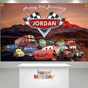 May include: A banner featuring the Cars movie characters with the text "Happy 3rd Birthday JORDAN". The background shows a desert landscape with mountains and a sunset. The banner has a white table in front of it with the words "Happy Birthday".