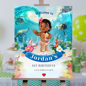 May include: Welcome to Jordan's 1st Birthday Celebration sign with a cartoon image of Moana, a tropical island scene, and a blue wave. The sign is on a wooden easel.