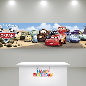 May include: A rectangular banner featuring a desert landscape with various animated car characters, including a red race car. The text reads Happy 3rd Birthday JORDAN. Below, a white table displays a colorful Happy Birthday sign in block letters.